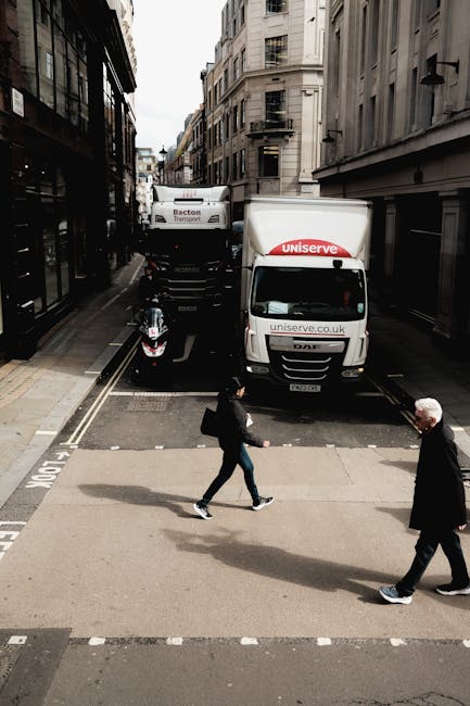 A city street scene showing a narrow, urban road lined with multi-story buildings featuring fire escapes and balconies, with traffic directing vehicles including a large white moving truck marked with the Uniserve logo and side panels, and a smaller van also branded with Uniserve for house removals and relocation services. The trucks are parked close to the curb, partially blocking the road, while a motorcycle is parked on the side pavement. In the foreground, two pedestrians are crossing the street; one person dressed in dark clothing with a backpack, walking briskly from left to right, and an older man with white hair and a black coat walking in the opposite direction. The lighting suggests overcast weather, with diffused natural light illuminating the scene. This image is related to packing, loading, and transportation tasks involved in home relocation services provided by Movers Harrow, reflecting the logistics of furniture transport and moving operations within an urban environment.