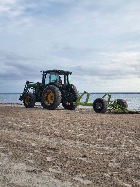 The image shows a large green tractor equipped with yellow wheels, situated on a sandy beach near the water's edge. Attached to the tractor is a long, lime-green implement with two large black tires, likely used for agricultural or ground-maintenance purposes. The tractor's cab is enclosed with glass windows, and the scene is under a partly cloudy sky, indicating daytime with natural light illuminating the area. In the background, the calm sea extends to the horizon, with no other vehicles or structures visible nearby. The setting appears quiet and open, with the tractor positioned slightly off-centre toward the left side of the image. Although the scene primarily depicts outdoor machinery usage, the image’s neutral, factual presentation can be related to equipment transportation or preparation activities in outdoor environments. This visual can be linked to logistical or transportation aspects of house removals and moving services, as provided by Movers Harrow, to support the topics of packing, loading, or equipment handling involved in home relocation processes.