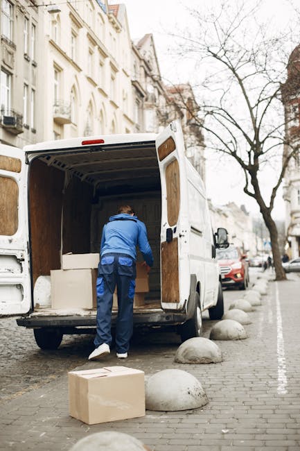 A city street scene showing a narrow, urban road lined with multi-story buildings featuring fire escapes and balconies, with traffic directing vehicles including a large white moving truck marked with the Uniserve logo and side panels, and a smaller van also branded with Uniserve for house removals and relocation services. The trucks are parked close to the curb, partially blocking the road, while a motorcycle is parked on the side pavement. In the foreground, two pedestrians are crossing the street; one person dressed in dark clothing with a backpack, walking briskly from left to right, and an older man with white hair and a black coat walking in the opposite direction. The lighting suggests overcast weather, with diffused natural light illuminating the scene. This image is related to packing, loading, and transportation tasks involved in home relocation services provided by Movers Harrow, reflecting the logistics of furniture transport and moving operations within an urban environment.