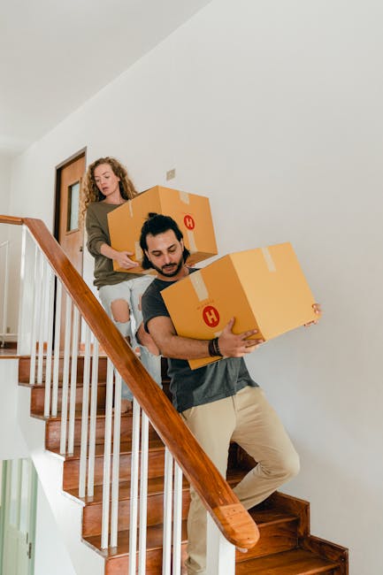 A man and a woman are carrying large cardboard boxes down a wooden staircase during a home relocation. The man is in front, holding a box close to his chest with both hands, wearing beige trousers and a dark t-shirt, focusing on the staircase. Behind him, a woman with curly hair, wearing a brown jacket and jeans with rips at the knees, is stepping down the stairs and passing a cardboard box to him. The staircase has wooden steps with a polished finish, a wooden handrail on the right, and white-painted balusters. The surrounding walls are painted light grey or white, and natural light illuminates the scene from an unseen window or entrance. This image exemplifies packing and moving activities typical of professional removals services, such as those provided by Movers Harrow, involving the careful handling and transportation of household items.