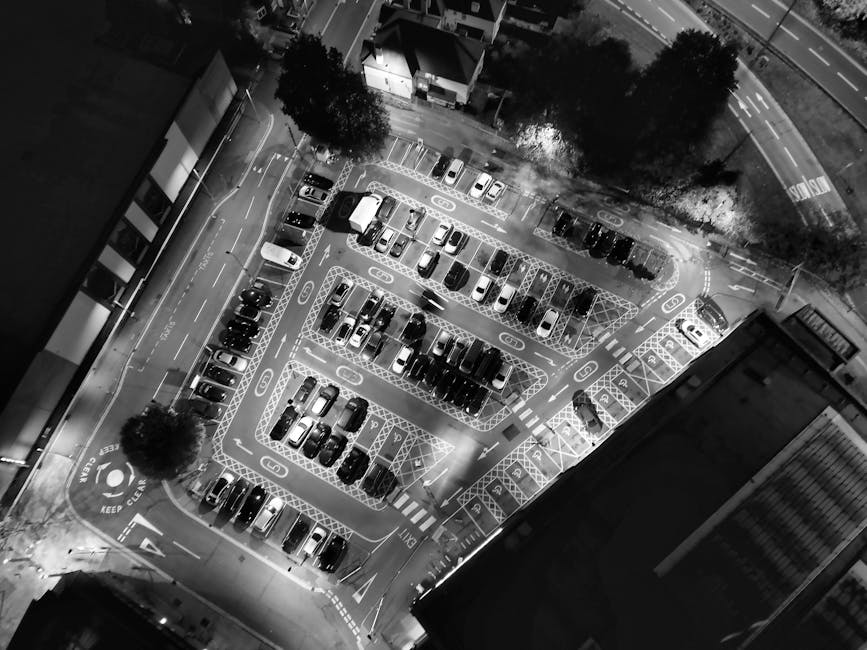 An overhead black-and-white image of a multi-storey car park situated on a city street at night. The parking lot is illuminated by artificial lighting and contains numerous parked vehicles, including cars and vans, arranged neatly within marked parking bays and designated loading zones. Several carts, likely used for moving boxes and furniture, are visible near the vehicles. Adjacent to the parking area, a building with large windows and a tree-lined sidewalk can be seen, along with road markings and traffic signs indicating parking restrictions and proper loading procedures. The scene captures the logistics of home relocation or furniture transport, with the structural layout supporting the moving process, and the environment emphasizing urban parking management as specified by Harrow Council parking rules for removals and loading, relevant to [PAGE_TITLE]. This visual is provided by Movers Harrow, illustrating typical parking conditions encountered during house removals in the area.