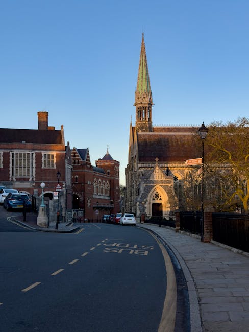 The image shows a narrow street in Harrow with a row of historic brick buildings on the left and a church with a tall, pointed spire on the right, both illuminated by natural daylight. The street has a bus stop marked 'BUS STOP' painted in white on the asphalt, and several parked cars are visible along the curb and pavement. In the foreground, a pair of trolleys, some packing boxes wrapped in plastic, and a wooden furniture dolly are positioned on the sidewalk, indicating recent or ongoing home relocation activities. A large cardboard box, protective blankets, and plastic wrapping materials are laid nearby, supporting furniture transport and packing during a move. The scene captures the logistical elements involved in house removals, with an emphasis on street parking regulations and loading practices near Harrow Council area.