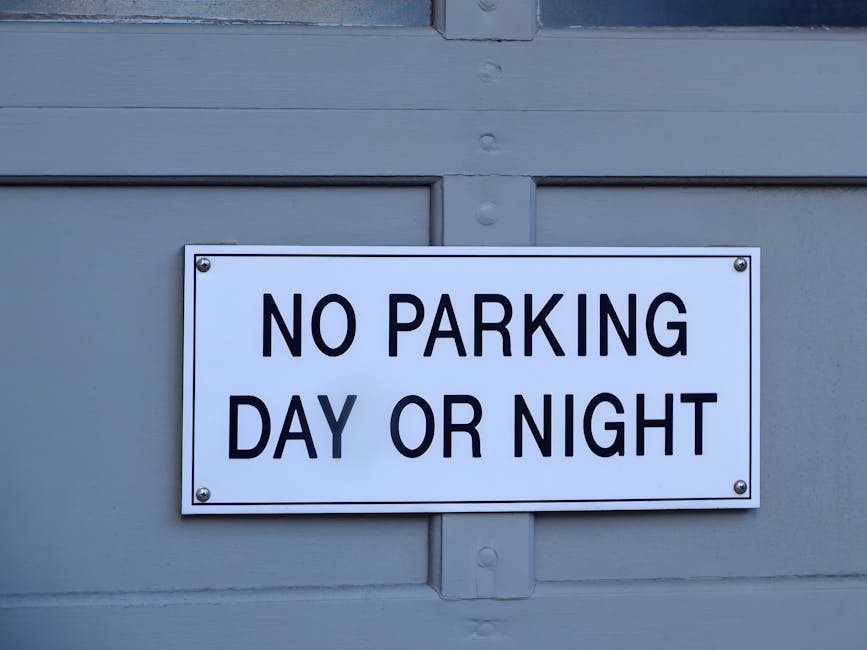 An overhead black-and-white image of a multi-storey car park situated on a city street at night. The parking lot is illuminated by artificial lighting and contains numerous parked vehicles, including cars and vans, arranged neatly within marked parking bays and designated loading zones. Several carts, likely used for moving boxes and furniture, are visible near the vehicles. Adjacent to the parking area, a building with large windows and a tree-lined sidewalk can be seen, along with road markings and traffic signs indicating parking restrictions and proper loading procedures. The scene captures the logistics of home relocation or furniture transport, with the structural layout supporting the moving process, and the environment emphasizing urban parking management as specified by Harrow Council parking rules for removals and loading, relevant to [PAGE_TITLE]. This visual is provided by Movers Harrow, illustrating typical parking conditions encountered during house removals in the area.