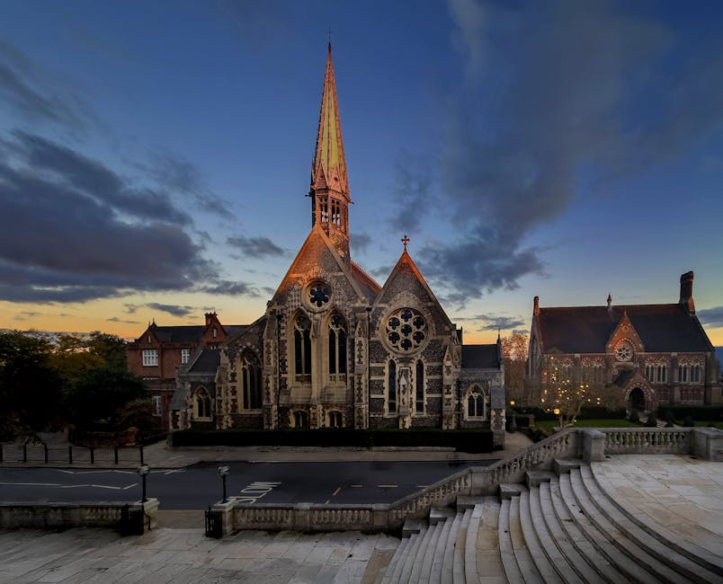A historic Gothic-style church with intricate stonework and large stained glass windows is illuminated by warm outdoor lighting at dusk, located within a small town or village setting. The church features a tall, pointed spire extending into the evening sky, with smaller gabled roof sections and detailed tracery on the windows. In front of the church, there is a paved area with a staircase leading down to the street, where lampposts line the sidewalk. To the right of the church, a neighboring building with similar architectural features is visible, also illuminated. The background consists of a partly cloudy sky transitioning from sunset to night, with soft clouds and fading light. The surroundings include some trees and residential buildings, and the scene captures a peaceful moment during the early evening. This image, associated with house removals and relocation services, underscores the importance of careful transportation logistics for preserving historic properties, as handled by Movers Harrow during home relocation projects.