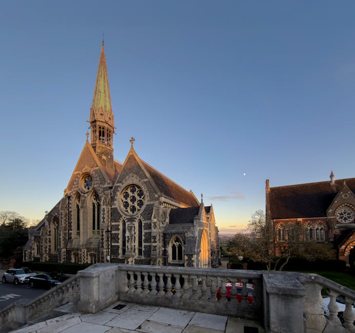 A historic Gothic-style church with intricate stonework and large stained glass windows is illuminated by warm outdoor lighting at dusk, located within a small town or village setting. The church features a tall, pointed spire extending into the evening sky, with smaller gabled roof sections and detailed tracery on the windows. In front of the church, there is a paved area with a staircase leading down to the street, where lampposts line the sidewalk. To the right of the church, a neighboring building with similar architectural features is visible, also illuminated. The background consists of a partly cloudy sky transitioning from sunset to night, with soft clouds and fading light. The surroundings include some trees and residential buildings, and the scene captures a peaceful moment during the early evening. This image, associated with house removals and relocation services, underscores the importance of careful transportation logistics for preserving historic properties, as handled by Movers Harrow during home relocation projects.