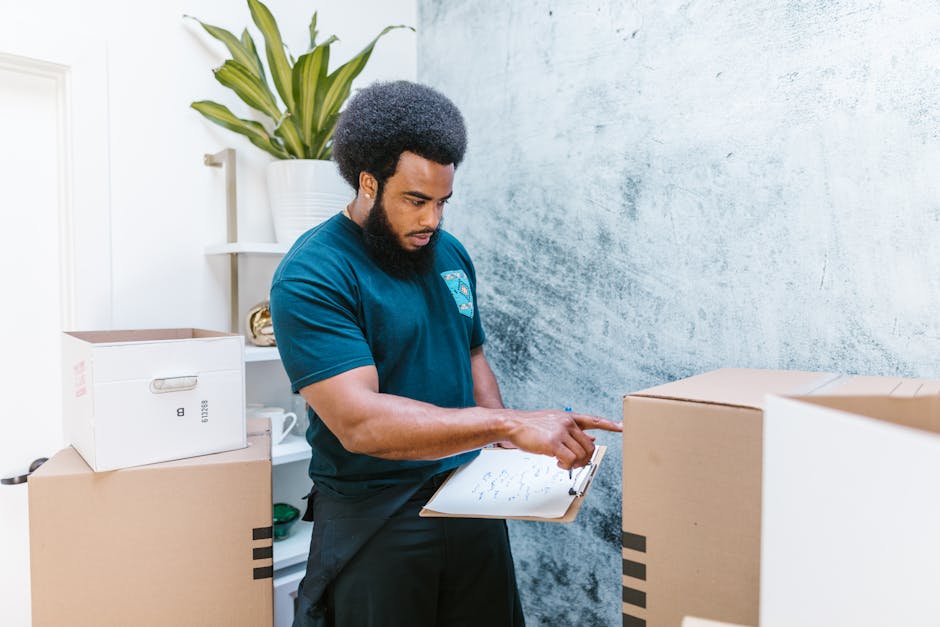 A professional removalist from Movers Harrow is inside a residential property, surrounded by several cardboard packing boxes of varying sizes, some sealed with packing tape and others open to reveal packing materials. The removalist is wearing a dark blue t-shirt and black trousers, holding a clipboard with a checklist and pointing at it with his index finger. The interior features a textured, light-coloured wall in the background, and a white shelving unit with a potted plant placed on top. The scene depicts the home relocation process, including packing preparations and organized site assessment, part of the furniture transport and loading operations associated with same-day Harrow removals, as the removalist prepares for the loading process with the boxes arranged around him for efficient transport by the company’s van, which is not visible in this image.