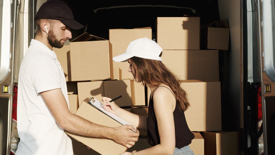 A man and a woman are engaged in a home relocation process inside a moving van, which is loaded with numerous cardboard boxes of various sizes, some covered with plastic wrap for protection. The man, dressed in a white polo shirt, holds a clipboard and appears to be checking or recording details, while the woman, wearing a white cap and black sleeveless top, writes on a notepad or forms attached to the clipboard. Both are positioned near the open rear doors of the van, which is parked on a paved area outside a property. The interior of the van is dimly lit, with natural light illuminating the scene from the open doors. The boxes are stacked neatly, some on dollies or hand trucks, indicating an ongoing loading process typical of furniture transport during house moving or packing and moving activities. This scene reflects professional removals by Movers Harrow, emphasizing the logistics of packing, loading, and home relocation services for same-day moves, as highlighted in the Last Minute Harrow Removals checklist page.