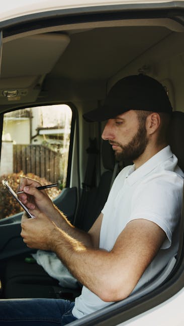 A professional removalist from Movers Harrow is inside a residential property, surrounded by several cardboard packing boxes of varying sizes, some sealed with packing tape and others open to reveal packing materials. The removalist is wearing a dark blue t-shirt and black trousers, holding a clipboard with a checklist and pointing at it with his index finger. The interior features a textured, light-coloured wall in the background, and a white shelving unit with a potted plant placed on top. The scene depicts the home relocation process, including packing preparations and organized site assessment, part of the furniture transport and loading operations associated with same-day Harrow removals, as the removalist prepares for the loading process with the boxes arranged around him for efficient transport by the company’s van, which is not visible in this image.