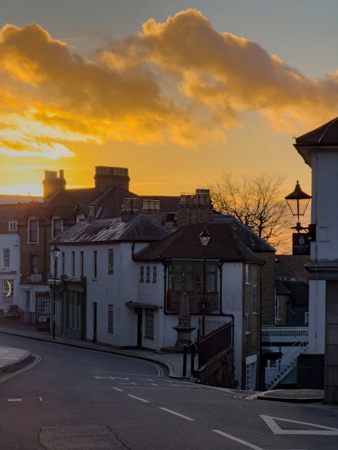 Photograph of a residential street during sunset, featuring a row of houses with various architectural styles, including brick and white-painted facades. Some houses have small front balconies, and a few have chimneys on their roofs. The street is paved with asphalt, showing road markings and mild curves, with a sidewalk on the left side. Street lamps line the pavement, and in the background, leafless trees and distant rooftops are visible against a sky filled with orange and yellow clouds, indicating the late evening atmosphere. The scene is well-lit by natural sunset light, highlighting the details of the buildings and street. This image depicts a quiet urban neighbourhood, relevant to home relocation and moving services, with potential for illustrating furniture transport, packing and moving operations, or the logistics involved in house removals in the Harrow area, as provided by Movers Harrow.