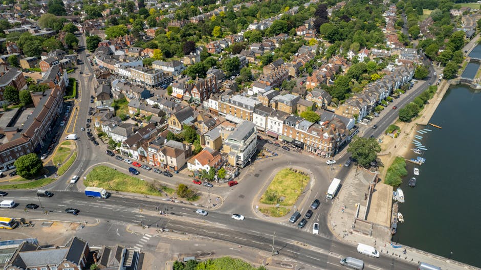 Aerial view of a town with a mix of residential and commercial buildings, including houses with pitched roofs, storefronts, and a harbor area with several small boats docked along the water's edge. The streets are busy with vehicles, including cars, trucks, and vans, some of which are parked along the roadside while others are in motion. There are open spaces and patches of greenery, with trees lining the roads and surrounding the buildings. The image shows a busy urban area possibly related to home relocation or moving services, with a waterfront setting and a mix of old and modern structures, as seen from above. This visual is suitable for a house removals and furniture transport context, emphasizing the logistical environment of moving and packing in Harrow, with the company Movers Harrow operating within this setting.
