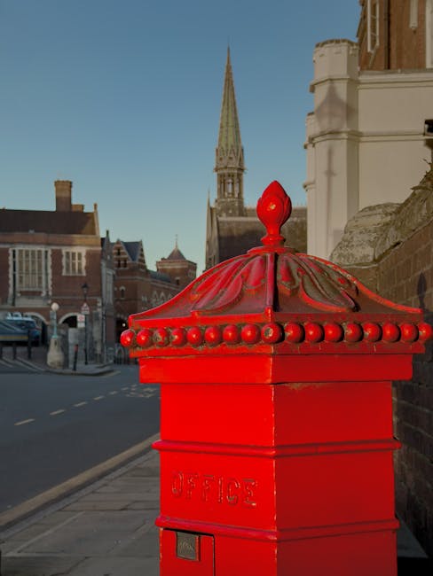 A street scene featuring a historic stone church with a tall, pointed spire topped with a green patina, situated next to a brick building with multiple windows. In the foreground, a paved road with double yellow lines runs uphill, with a red car parked on the left side and a white car parked near the church entrance on the right. There are black metal bollards along the sidewalk and a vintage-style street lamp mounted on a stone pillar near the church steps. The sky overhead is partly cloudy with patches of blue visible, and lush green trees are seen in the background. This scene could relate to house removals or home relocation services by Movers Harrow, highlighting the importance of careful transport and logistics during the moving process.
