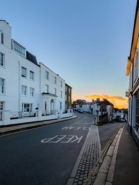 A quiet residential street in Harrow during sunset, with white multi-storey buildings on the left and mixed-height houses on the right. The road has a 'KEEP CLEAR' marking painted in white, and a narrow sidewalk with a brick-paved edge runs along the right side of the street. The street curves gently uphill, with a vanishing point toward the horizon where the sky displays warm orange and yellow hues blending into blue. While no furniture or packaging is visible in the image, a moving company such as Movers Harrow would typically set up packing materials like cardboard boxes, bubble wrap, and blankets nearby for transporting household items during a home relocation or furniture transport process. The scene captures an ideal setting for loading or unloading belongings from a removals van, with natural daylight illuminating the area.
