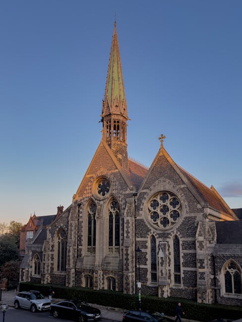 A wide view of a narrow street in Harrow on the Hill during daylight with clear skies, showing parked cars along the pavement, a traffic bus lane marked on the road, and a historic church with a tall, pointed spire and stained glass windows at the end of the street. Nearby buildings include brick structures with one featuring large sash windows, and a lamppost situated near the sidewalk. Visible in the scene are moving boxes, packing materials, and furniture covered with blankets, indicating a home relocation process managed by Movers Harrow. The image captures the typical urban setting for furniture transport and the logistical planning needed for house removals in tricky street layouts, emphasizing the importance of careful loading and transport within residential areas.