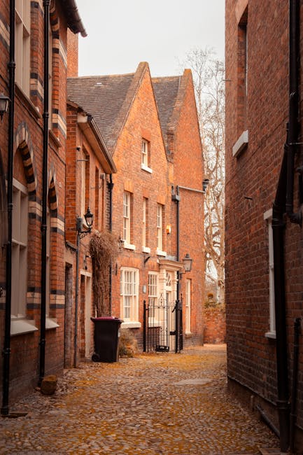 A narrow cobbled alleyway between red-brick residential buildings with small windows and white window frames. The alley is lined with a black metal gate at the far end, with a black trash bin positioned near the left side and a small lantern-style exterior light mounted on the building wall. The scene shows an outdoor setting during daylight hours with overcast skies, typical of a residential area in Wealdstone, Harrow. This environment depicts a typical home relocation or moving process in an area with limited access, relevant to house removals and furniture transport services offered by Movers Harrow. The detailed view of the brickwork, cobbled surface, and limited access space emphasizes the logistical considerations involved in narrow access removals, highlighting the importance of careful packing and loading activities during a house move.